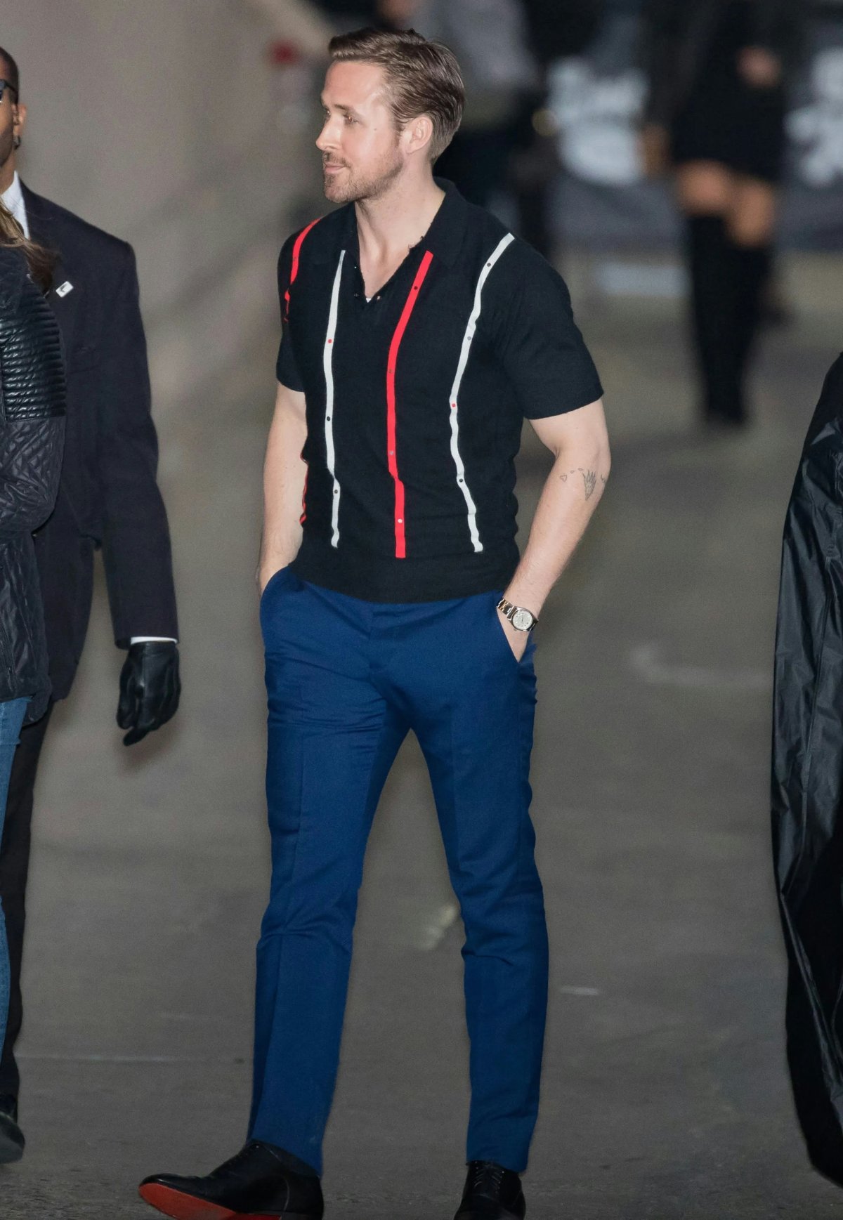 Ryan Gosling in a blue pants, black polo, on a brown background.